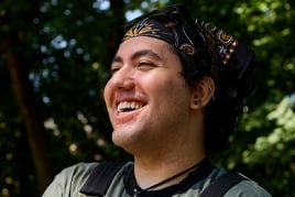 A smiling man wearing a decorated black hat outdoors among green trees