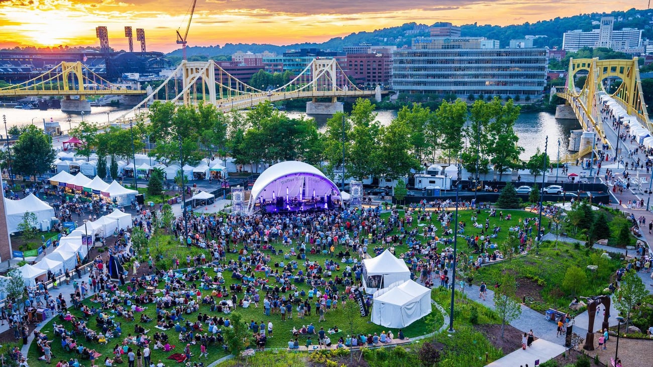 A large crowd gathers around an outdoor stage for a concert in a riverside park, with yellow bridges, city buildings, and a sunset in the background. White tents and lush greenery surround the event area.