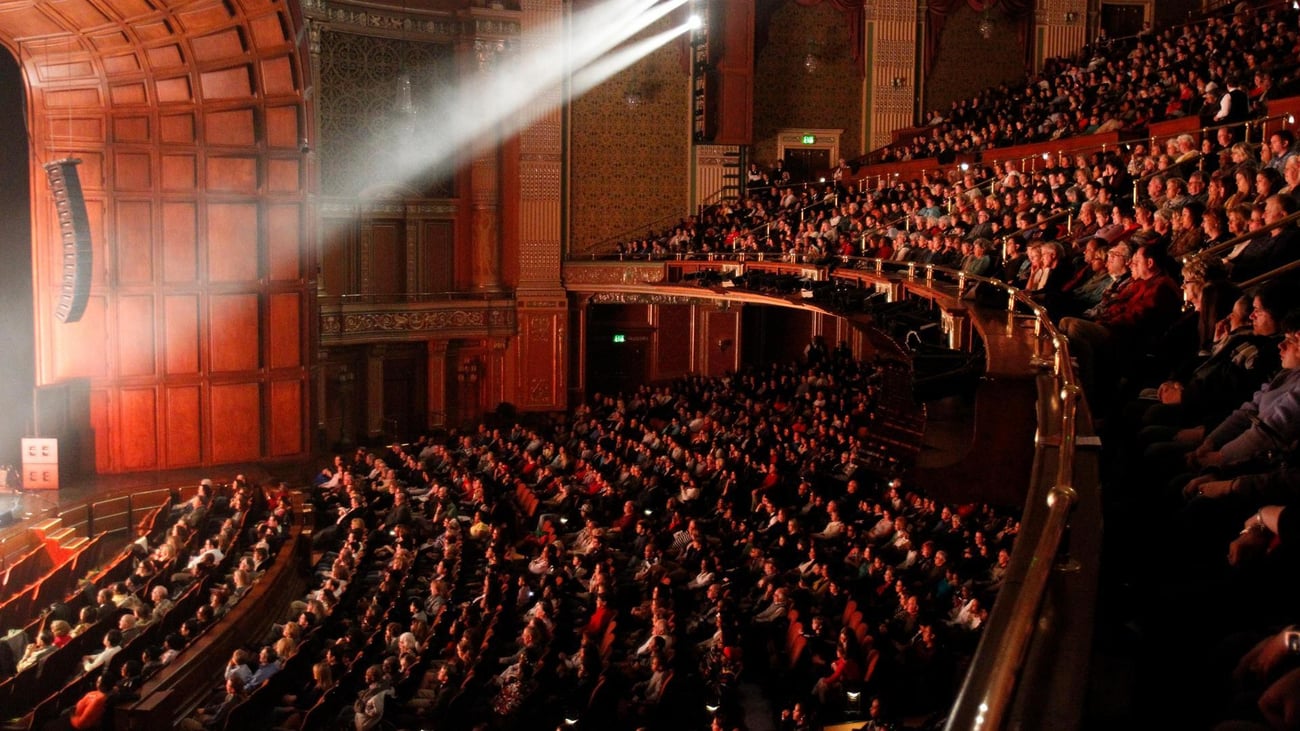 A large audience fills the seats of an ornate theater, watching a performance on stage. Warm lighting and a spotlight beam illuminate the elegant interior and attentive crowd. A large audience fills the seats of an ornate theater, watching a performance on stage. Warm lighting and a spotlight beam illuminate the elegant interior and attentive crowd.