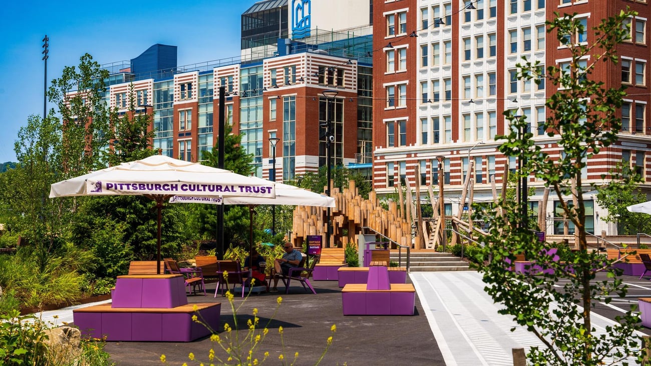 Outdoor urban park with purple benches, wooden play structures, and umbrellas labeled “Pittsburgh Cultural Trust.” People are sitting and relaxing, with modern red-brick buildings in the background. Outdoor urban park with purple benches, wooden play structures, and umbrellas labeled “Pittsburgh Cultural Trust.” People are sitting and relaxing, with modern red-brick buildings in the background.