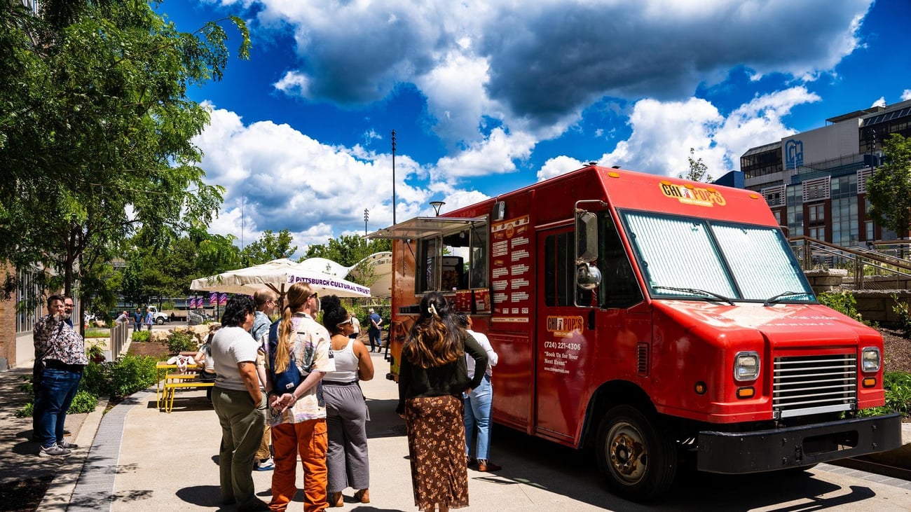 A group of people waits in line at a bright red food truck parked on a sunny sidewalk, with trees, umbrellas, and buildings in the background under a blue sky with scattered clouds. A group of people waits in line at a bright red food truck parked on a sunny sidewalk, with trees, umbrellas, and buildings in the background under a blue sky with scattered clouds.