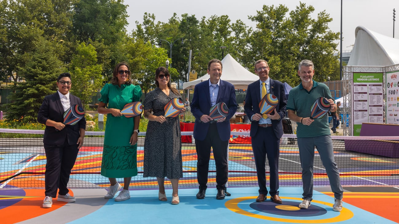 Six people stand on a colorful outdoor court, holding rackets and smiling for the camera, with trees and a tent in the background.