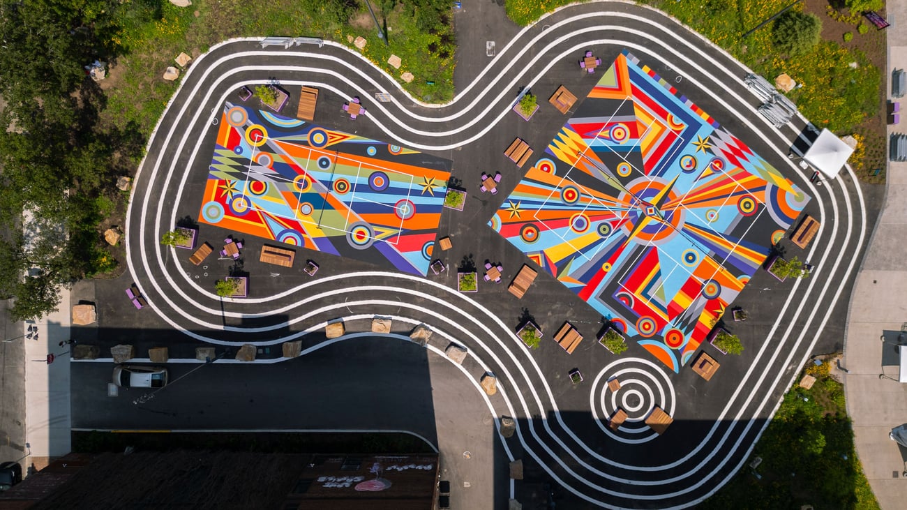 Aerial view of a vibrant public space with colorful geometric murals, winding white lines, benches, tables, and people sitting or walking, surrounded by greenery and urban elements. Aerial view of a vibrant public space with colorful geometric murals, winding white lines, benches, tables, and people sitting or walking, surrounded by greenery and urban elements.