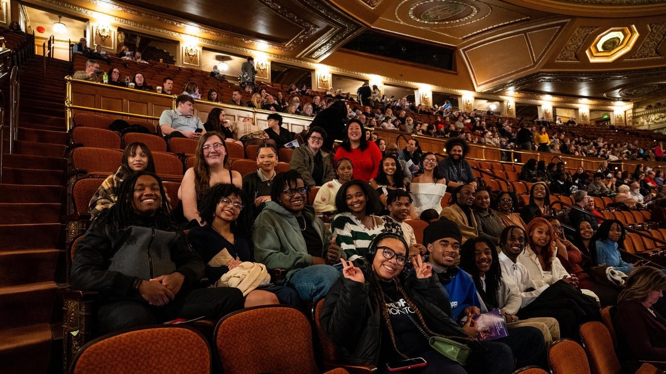 A diverse group of people smile and pose for a photo inside a large, ornate theater with many others seated in the background. A diverse group of people smile and pose for a photo inside a large, ornate theater with many others seated in the background.