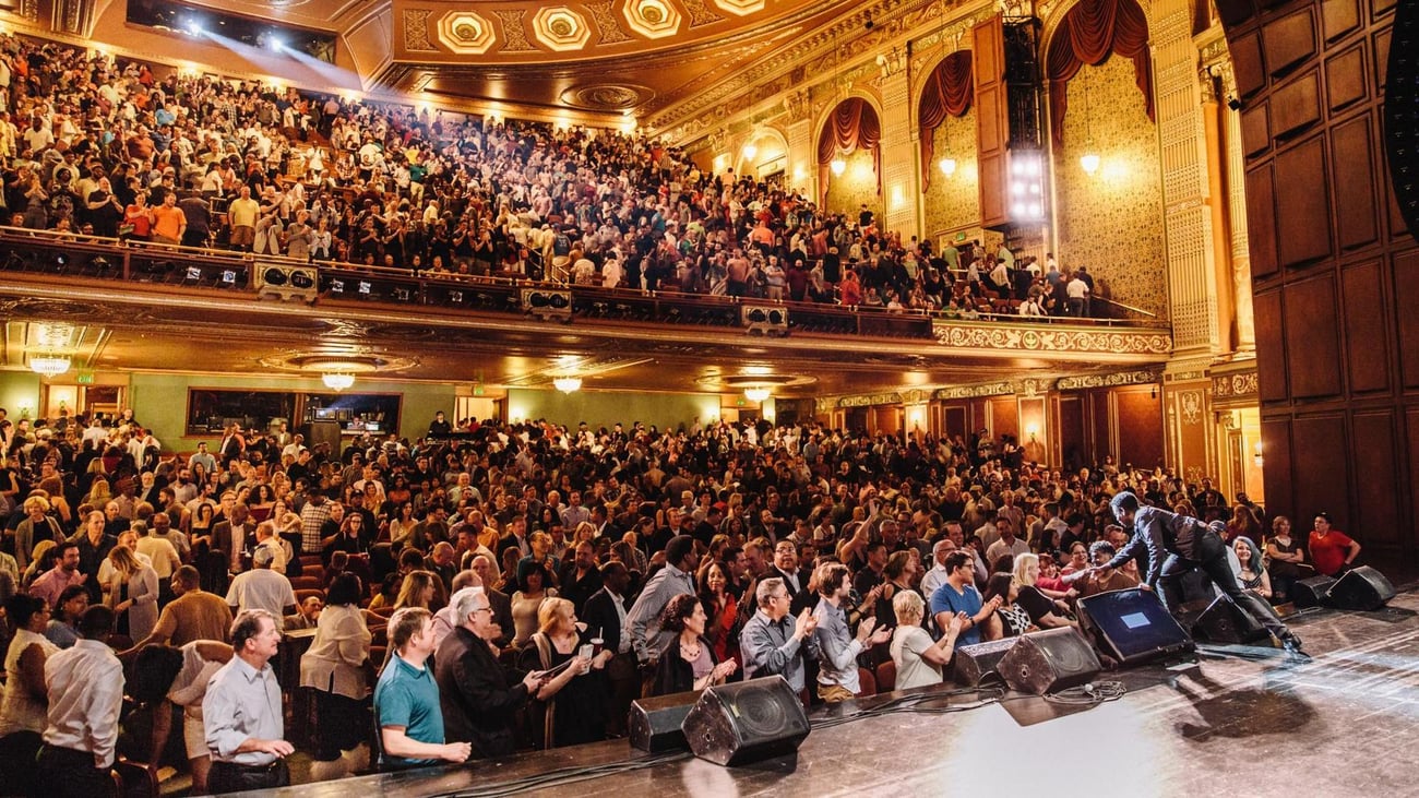 A performer on stage greets a large, cheering audience in an ornate, packed theater with golden decor and balcony seating.