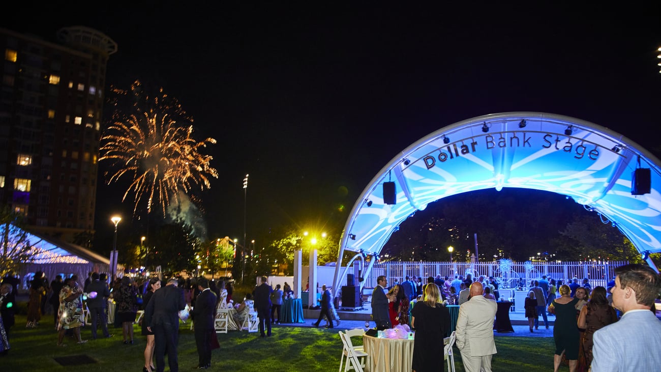 People gather at night near an outdoor stage labeled Dollar Bank Stage, with fireworks lighting up the sky in the background. Some guests are mingling while others watch the performance on the illuminated stage.