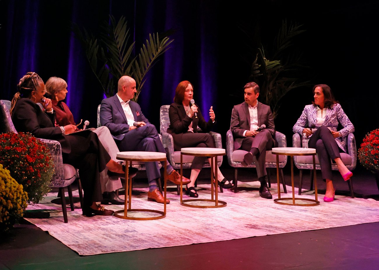 Six people in business attire sit on stage in armchairs, engaged in a panel discussion. One woman speaks while others listen. The background features plants and blue-purple lighting. Small tables and red flowers are in front of them. Six people in business attire sit on stage in armchairs, engaged in a panel discussion. One woman speaks while others listen. The background features plants and blue-purple lighting. Small tables and red flowers are in front of them.
