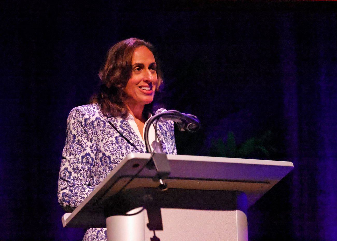 A person in a blue patterned jacket speaks at a podium on a dimly lit stage, illuminated by a spotlight. A person in a blue patterned jacket speaks at a podium on a dimly lit stage, illuminated by a spotlight.