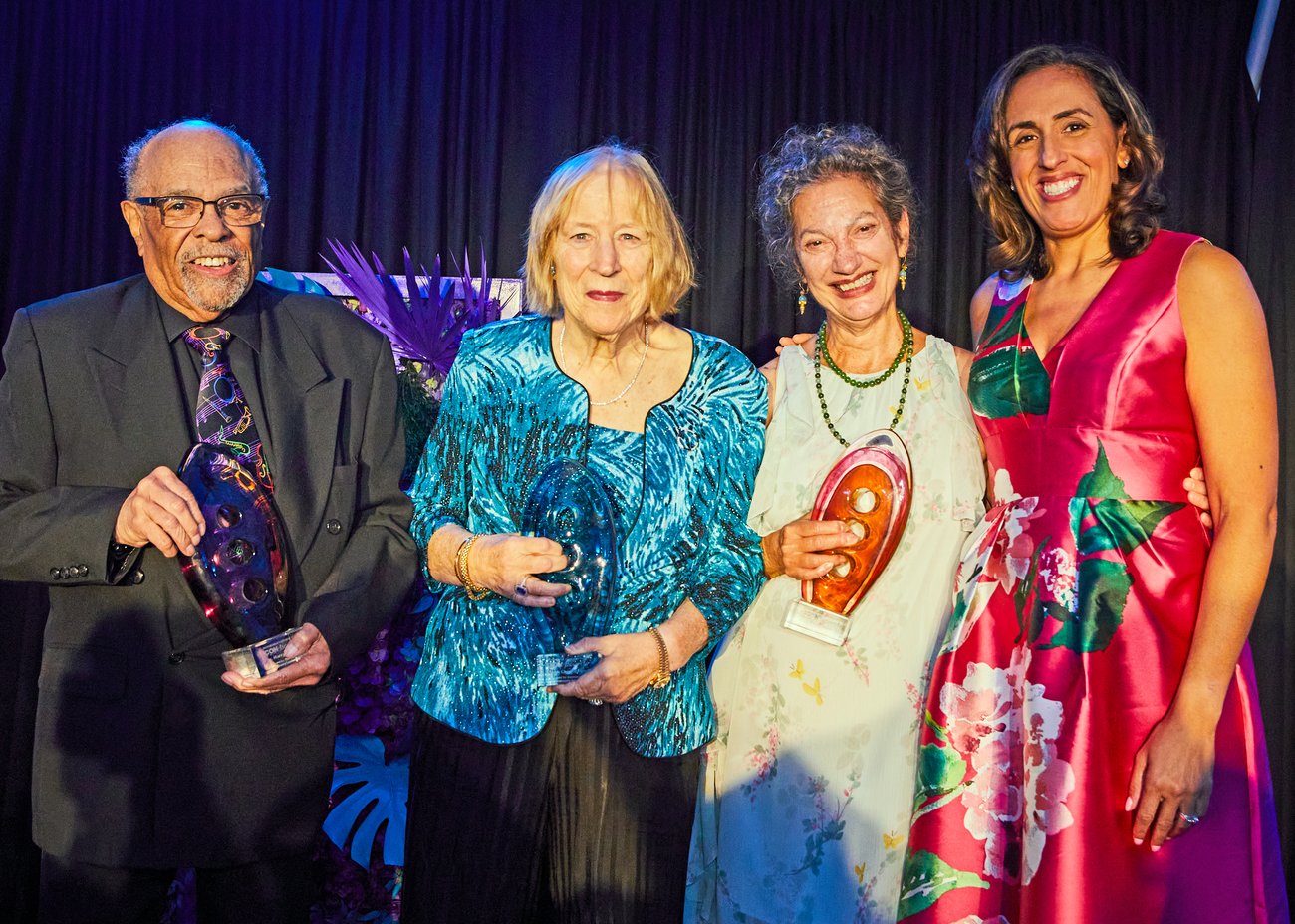 Four adults stand together smiling, three holding glass awards. They are dressed in formal attire with a dark curtain and floral display in the background. Four adults stand together smiling, three holding glass awards. They are dressed in formal attire with a dark curtain and floral display in the background.