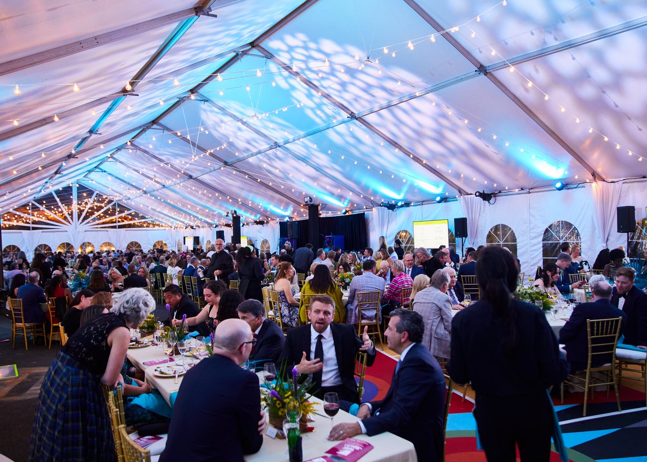 A large group of people in formal attire dine and mingle under a festively lit white tent with string lights and colorful decor. A large group of people in formal attire dine and mingle under a festively lit white tent with string lights and colorful decor.