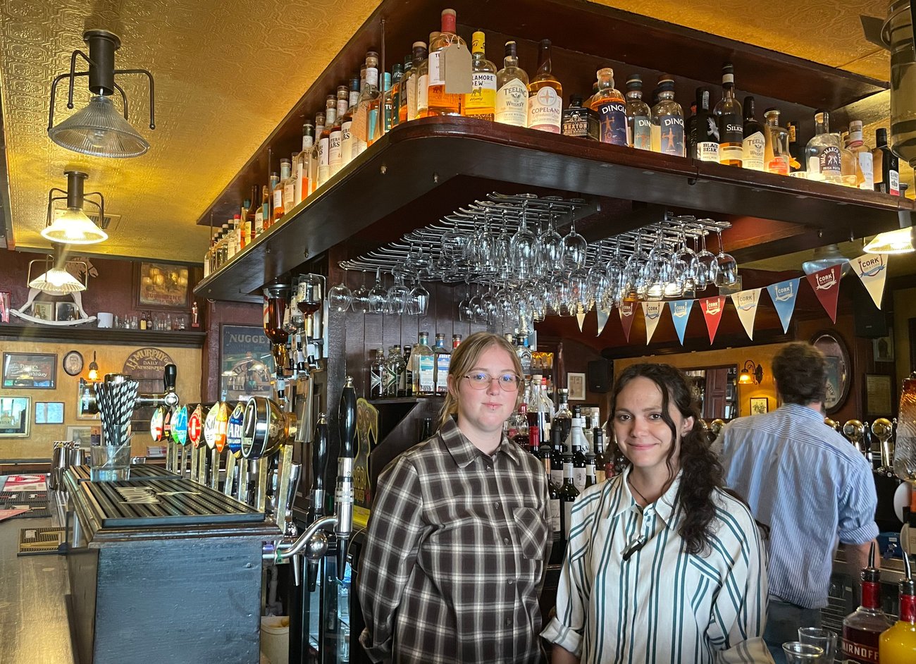 Two people pose behind a pub bar surrounded by bottles and hanging glasses. Two people pose behind a pub bar surrounded by bottles and hanging glasses.