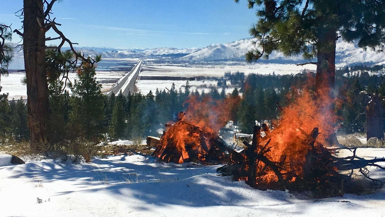 A scenic, high-angle landscape photograph showing active slash pile burning on a snowy hillside overlooking a vast mountain valley. The Foreground: On a snow-covered slope, two large piles of cut logs, branches, and woody debris are burning with bright orange and red flames. Light blue smoke rises from the fires. A few evergreen trees frame the left and right sides of the immediate foreground. The Background Landscape: Below the hillside, a dense green forest of pine trees leads down to a wide, flat valley floor blanketed in white snow. A long, straight multi-lane highway stretches directly through the center of the valley toward the horizon. The Horizon: In the far distance, a massive range of rolling mountains covered in snow sits under a clear, bright blue sky.