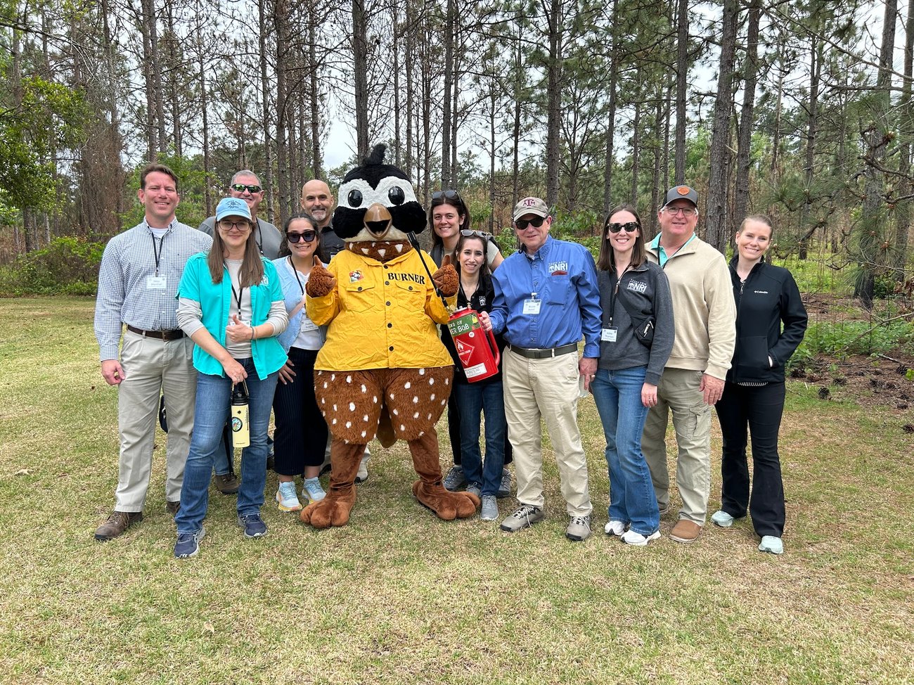 A wide, eye-level outdoor group photograph of ten people standing on a grassy lawn alongside Burner Bob in front of a pine forest under an overcast sky.