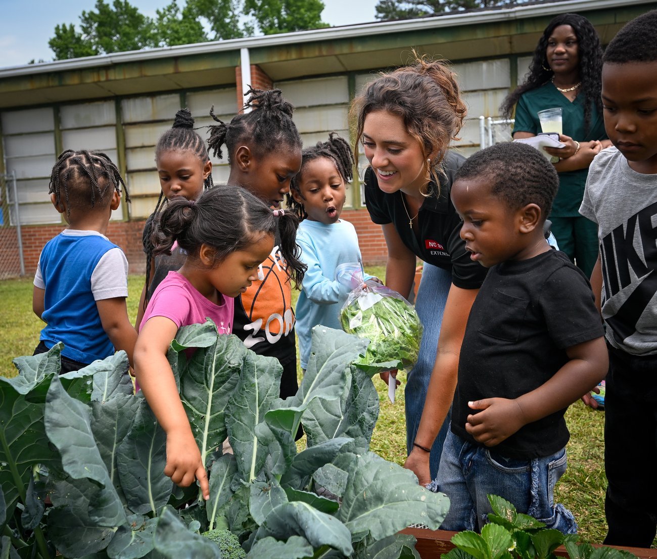 An NC State Extension staff member smiles as she shows a head of broccoli to a group of young children gathered around a raised garden bed outdoors.