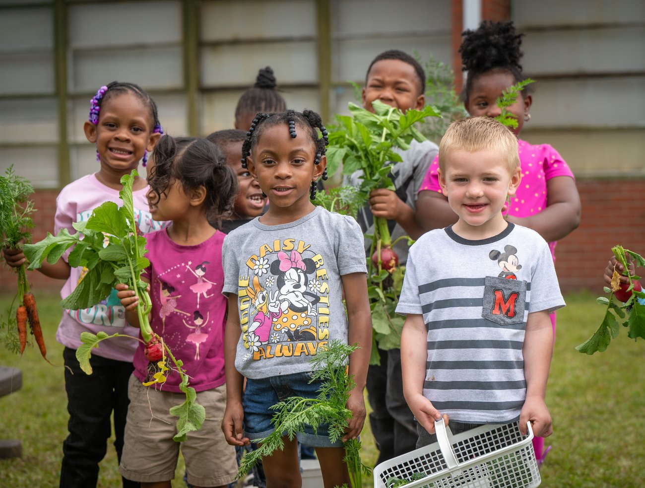 A group of young children smile and hold freshly harvested vegetables, including carrots and radishes, outside their early care center.