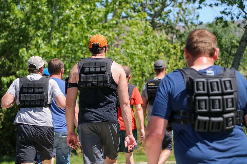 A view from behind of a group of men walking outdoors on a sunny day, taking part in a pack test. Several of the men are wearing heavy, black weighted vests over their t-shirts and tank tops. Lush green trees and foliage line the path ahead of them under a clear blue sky.