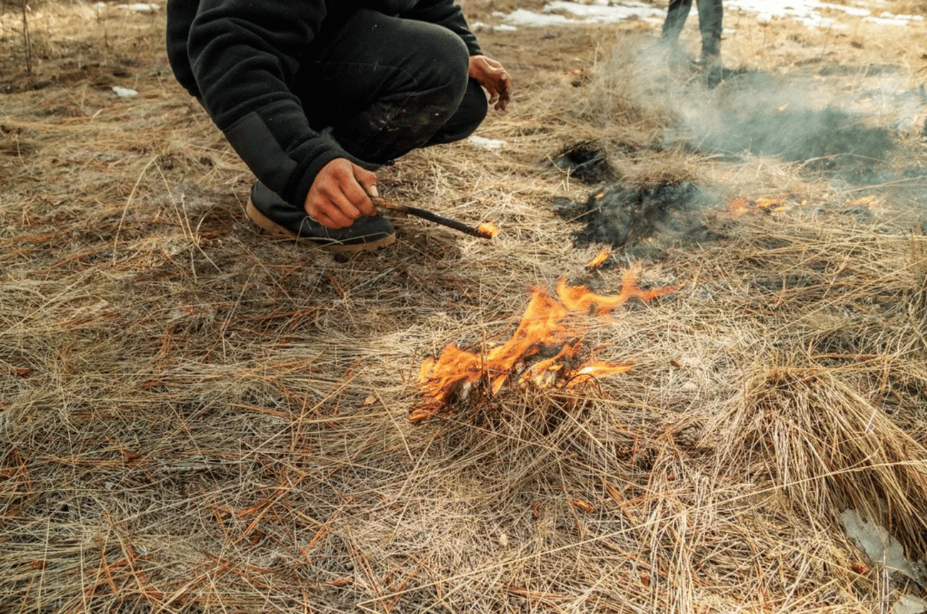 A close-up, low-angle shot of a person crouching down in a field of dry, brown grass. The person is using a small, burning stick held in their hand to deliberately ignite a small patch of grass, creating bright orange flames. In the upper right background, a small patch of scorched earth produces wisps of smoke, and the legs of another person can be seen standing nearby.