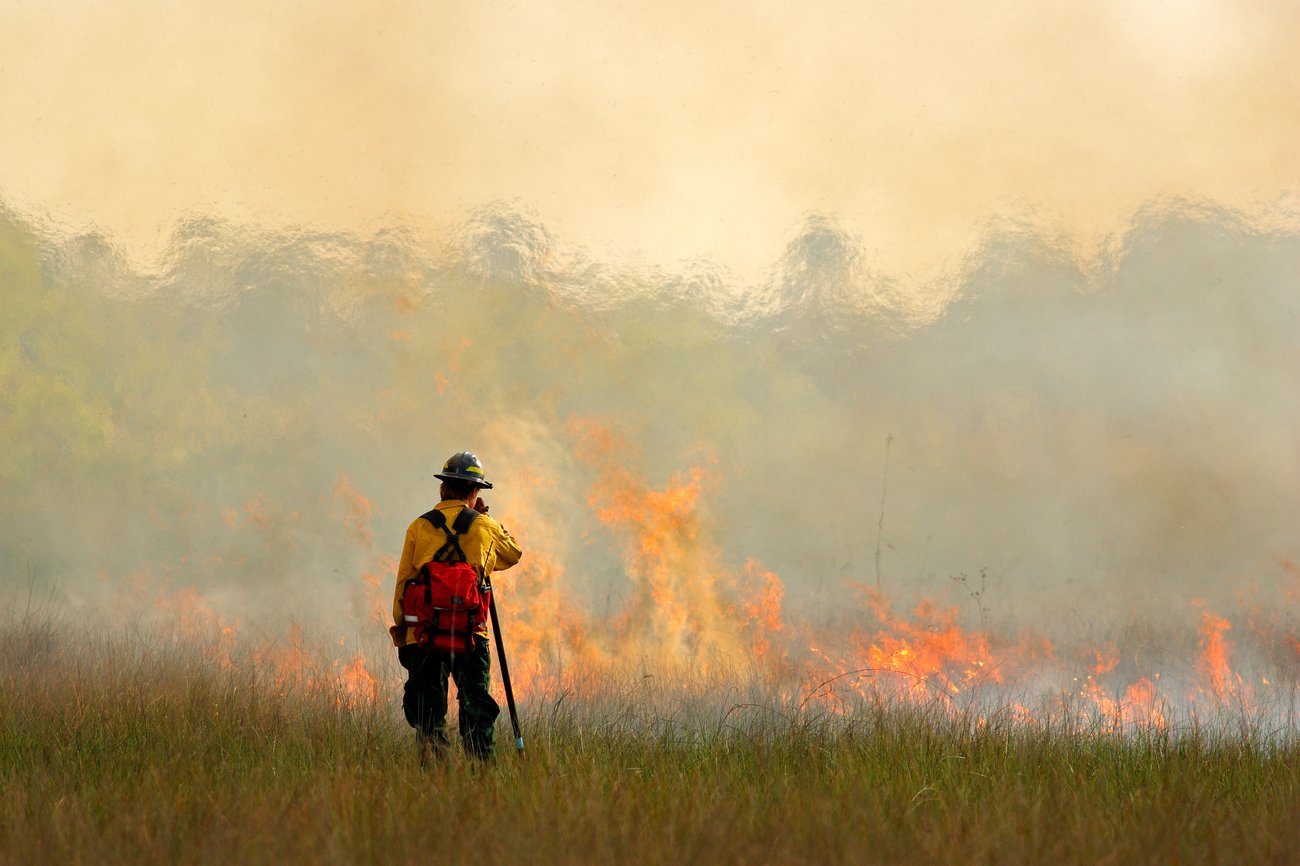 A wildland firefighter stands in a grassy field with their back to the camera, monitoring a line of bright orange flames and thick smoke sweeping across the landscape. The firefighter wears a yellow fire-resistant jacket, dark green pants, a hard hat, and a prominent red backpack while holding a tool or radio. In the background, intense heat waves blur the upper horizon against a hazy, smoke-filled sky.