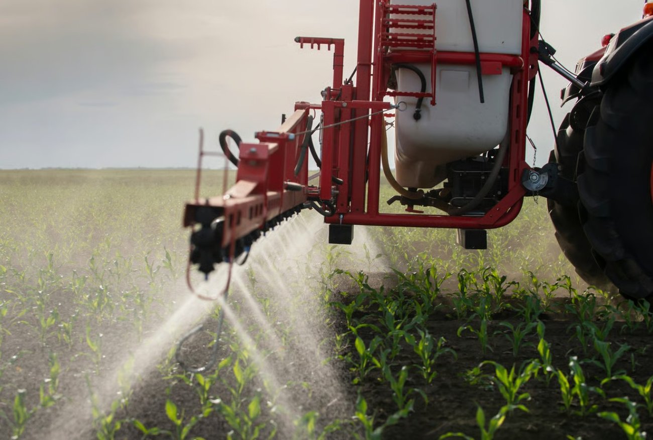 A tractor driving through a farm field while spraying lines of liquid pesticide over rows of green crops.