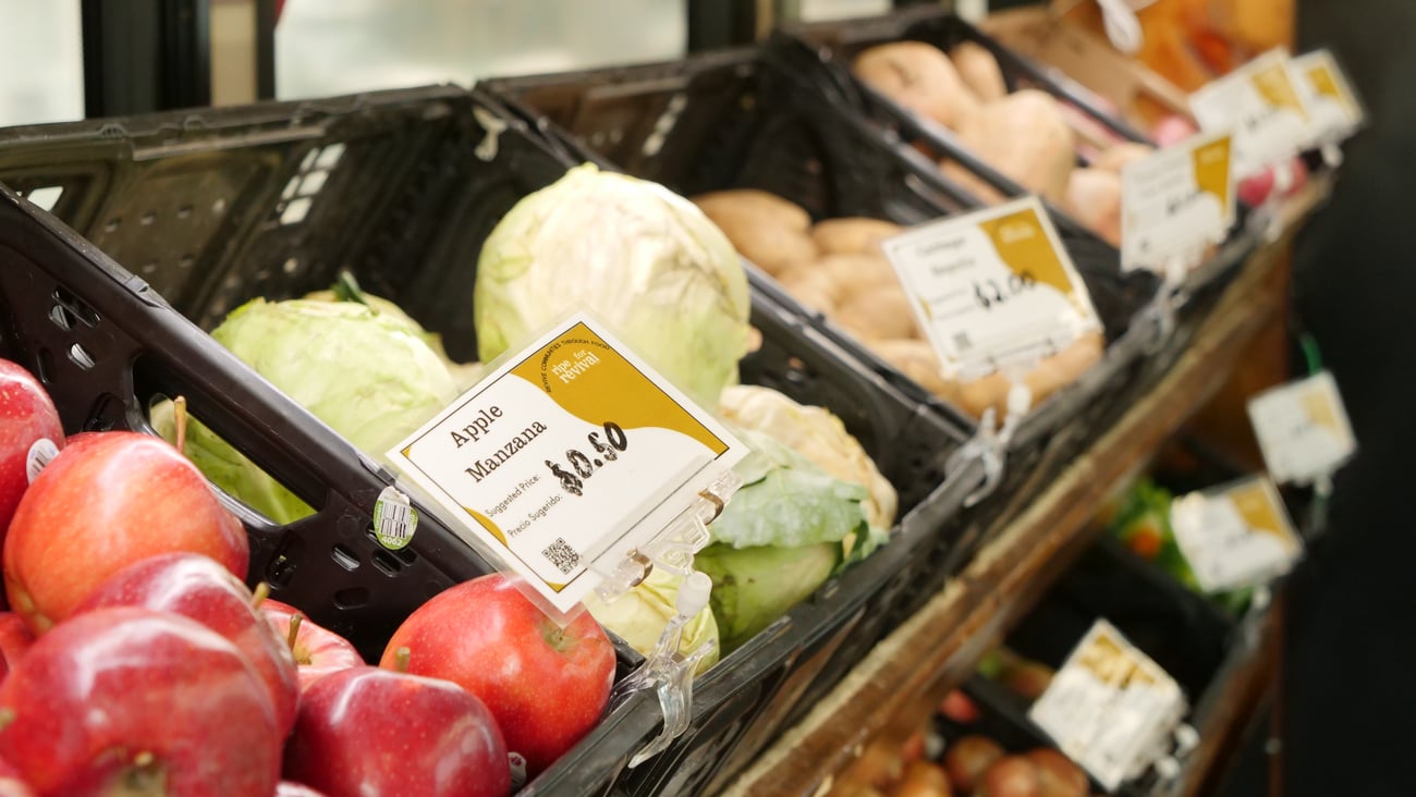 Black produce bins display fresh fruits and vegetables, including red apples, green cabbage, and potatoes, with bilingual English and Spanish price signs at a mobile market.