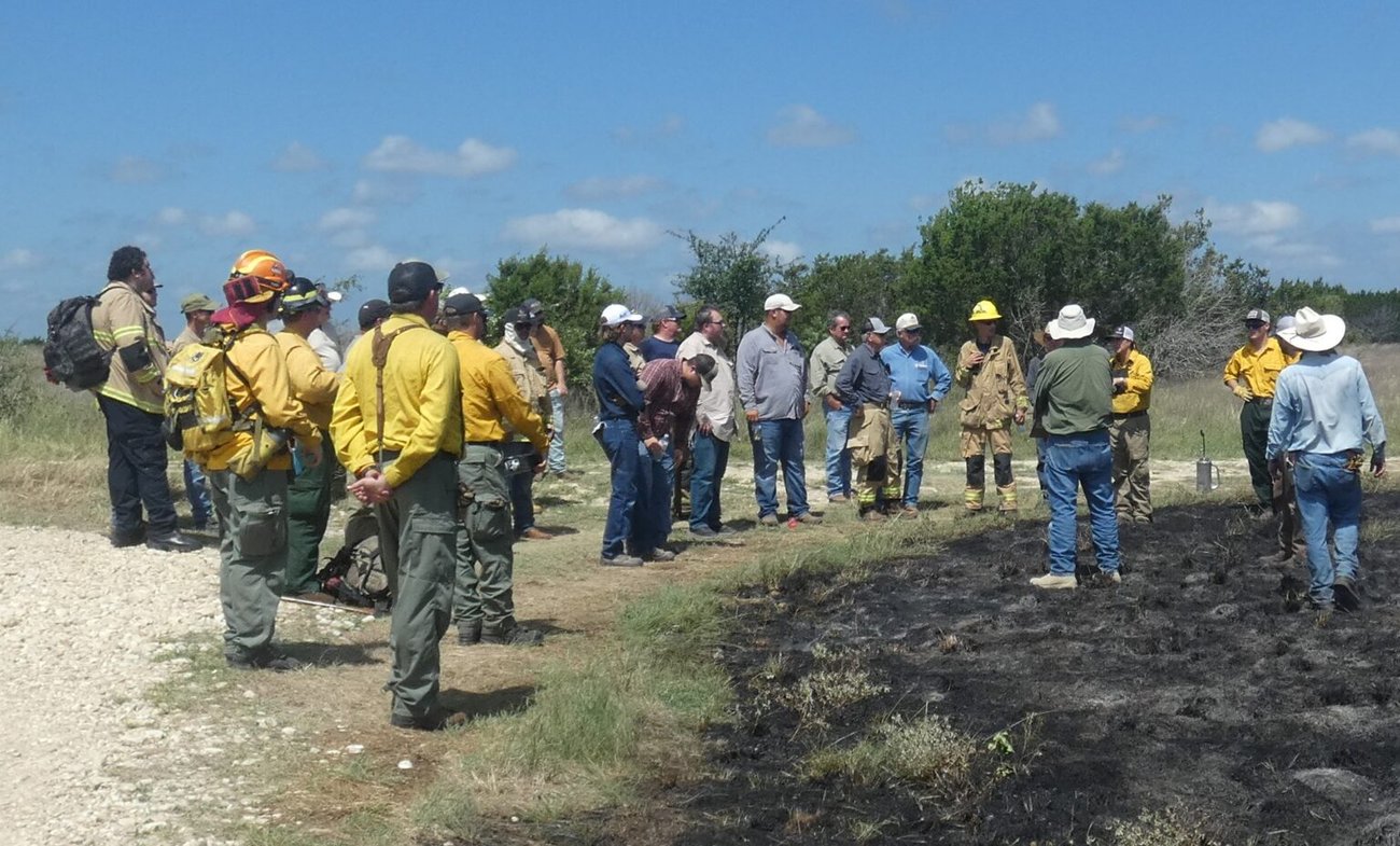 A large group of natural resource professionals, wildland firefighters, and local landowners gathered outdoors next to a freshly burned patch of ground. They are wearing yellow Nomex field shirts, hard hats, and casual work attire while standing in a circle along a dirt road during a field demonstration.