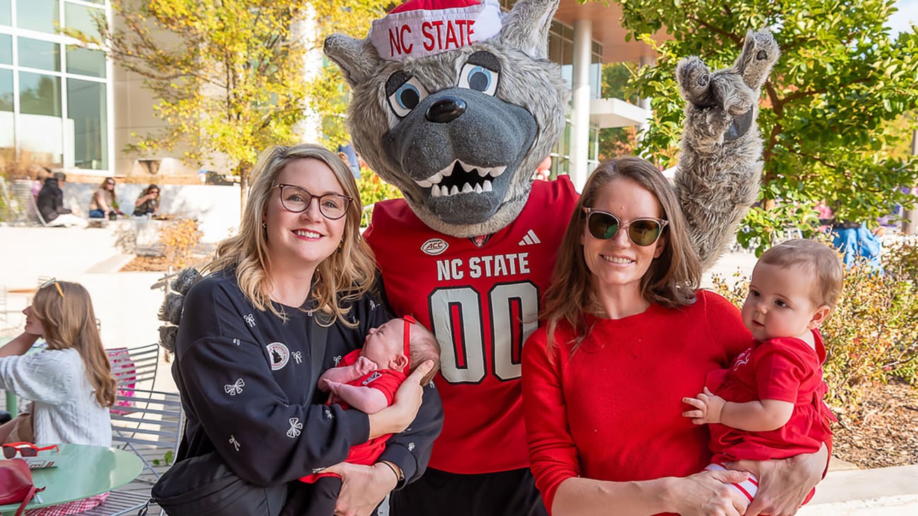 NC State male mascot Mr. Wuf stands outside with two women holding their babies during the 2025 CALS Wolfpack Harvest celebration.