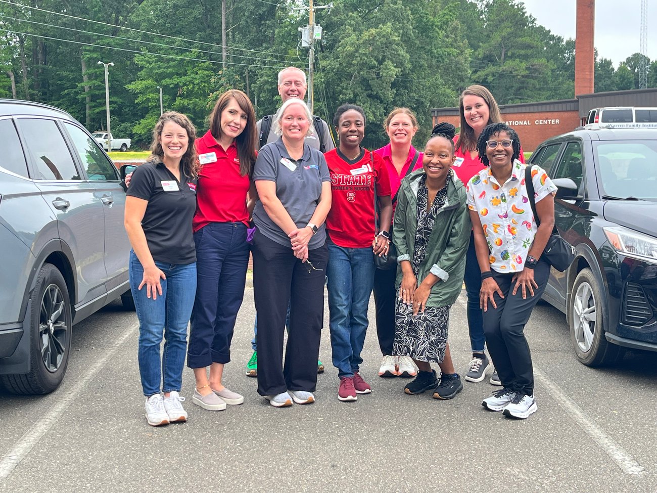 Nine Healthy ROOTS team members and partners smile together in a parking lot outside a community center. Team members wear a mix of red NC State polos, casual attire, and name badges. Trees and a brick building are visible in the background.