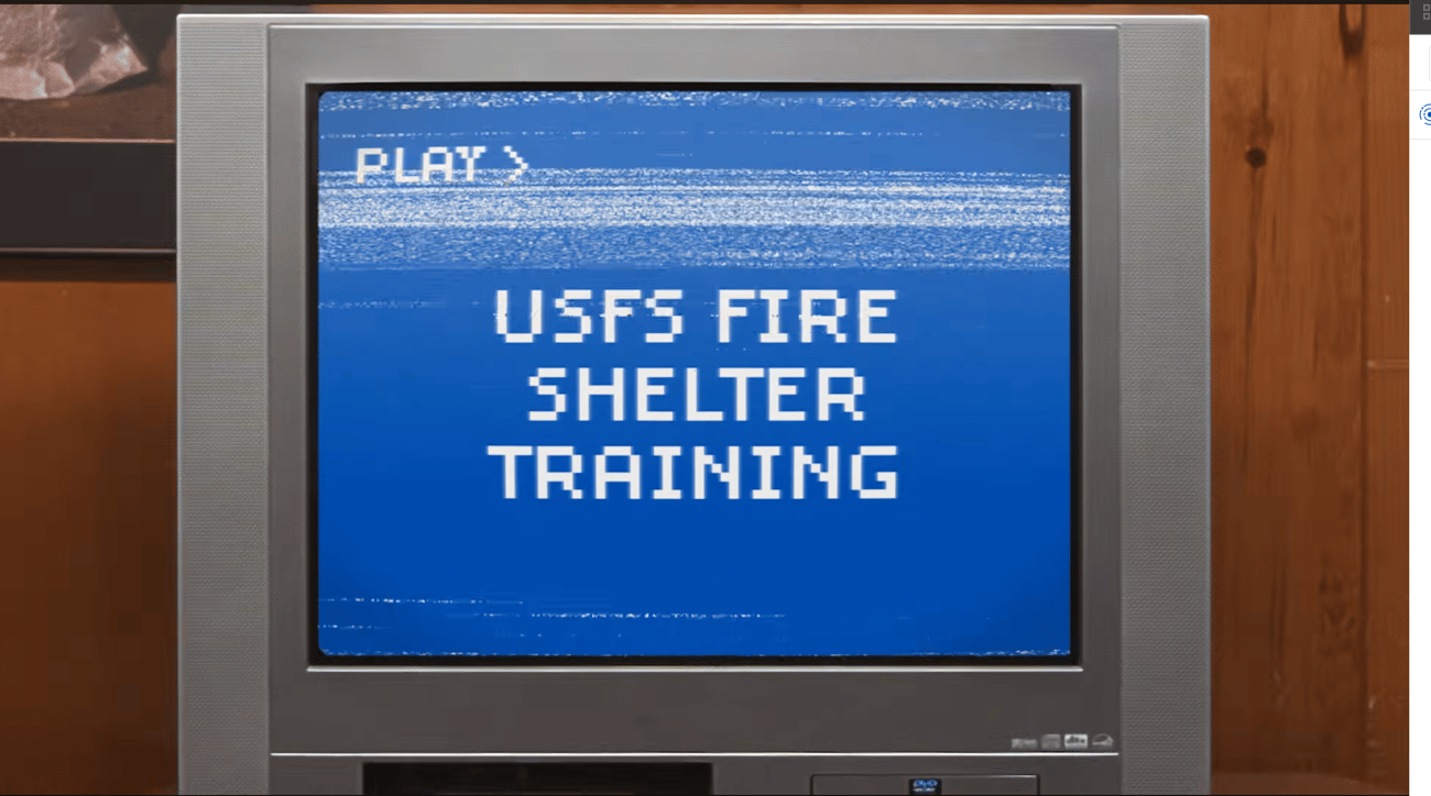 An old-school silver television set set against a wood-paneled wall. The TV screen shows static and VHS-style tracking distortion over a blue background featuring white, pixelated retro text reading "PLAY > USFS FIRE SHELTER TRAINING."