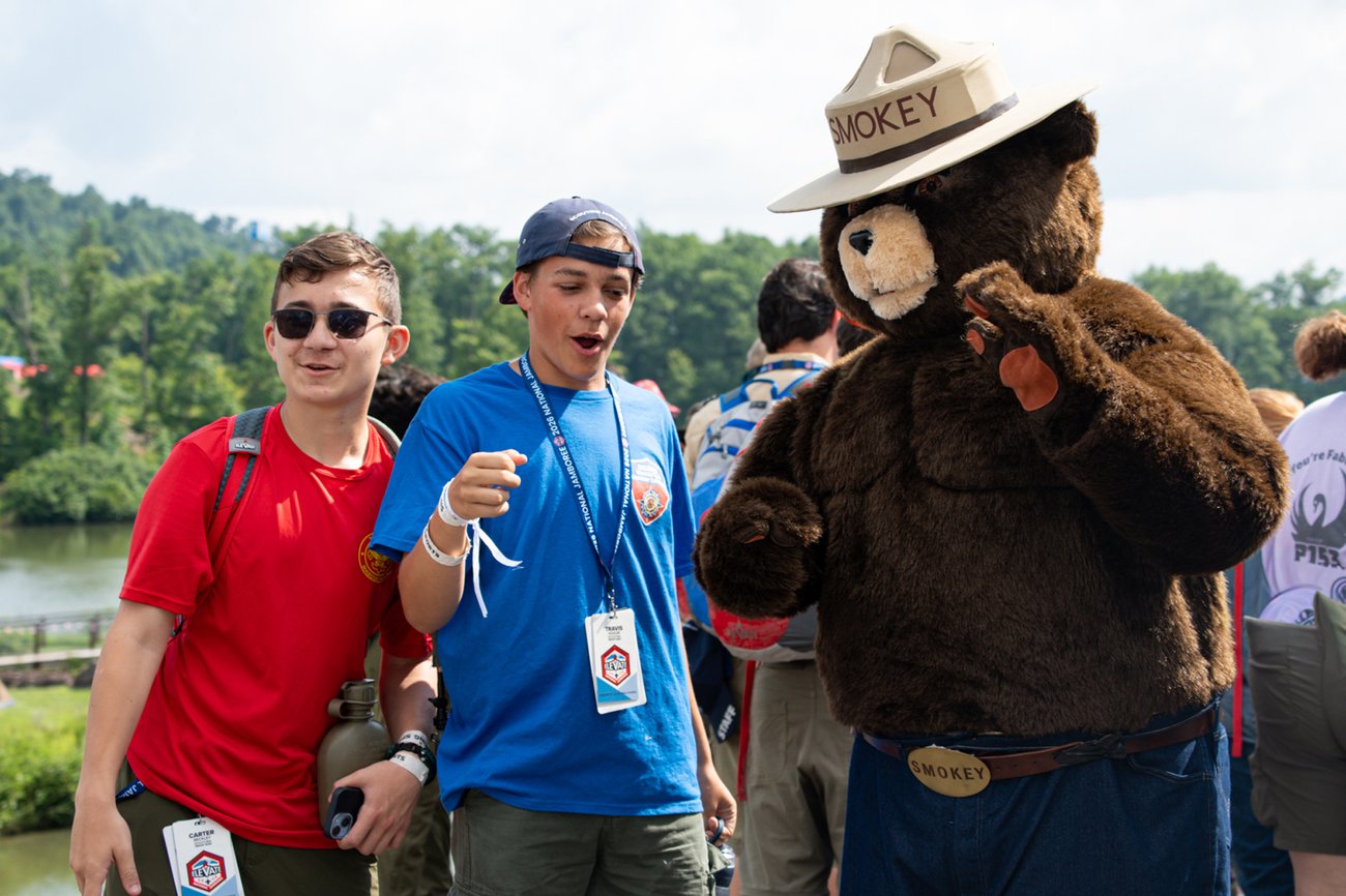 Two youth wearing Scouting America lanyards interact outdoors with a Smokey Bear as he wears his classic campaign hat and belt buckle. The scout in the blue shirt reacts with excitement while Smokey Bear waves his paw.