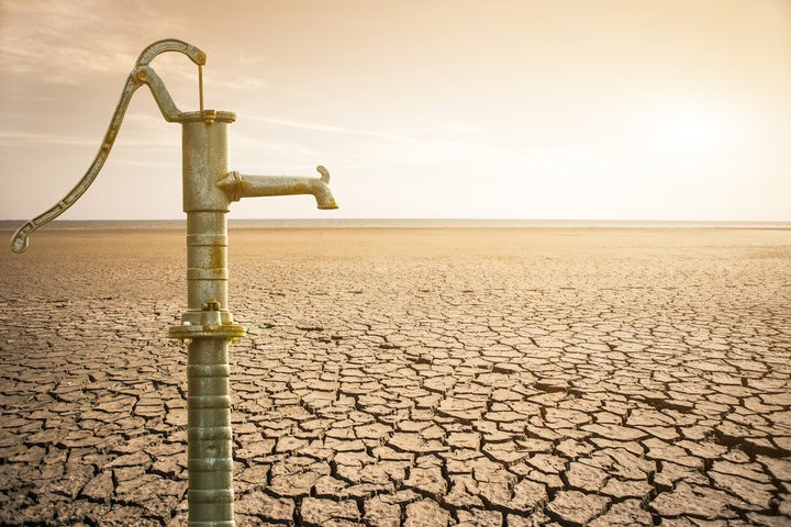 A manual well pump in the foreground, in the background an expanse of dry, parched earth.