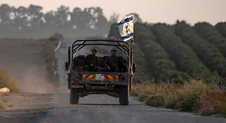 Two men ride in the back of a truck on a dusty rural road