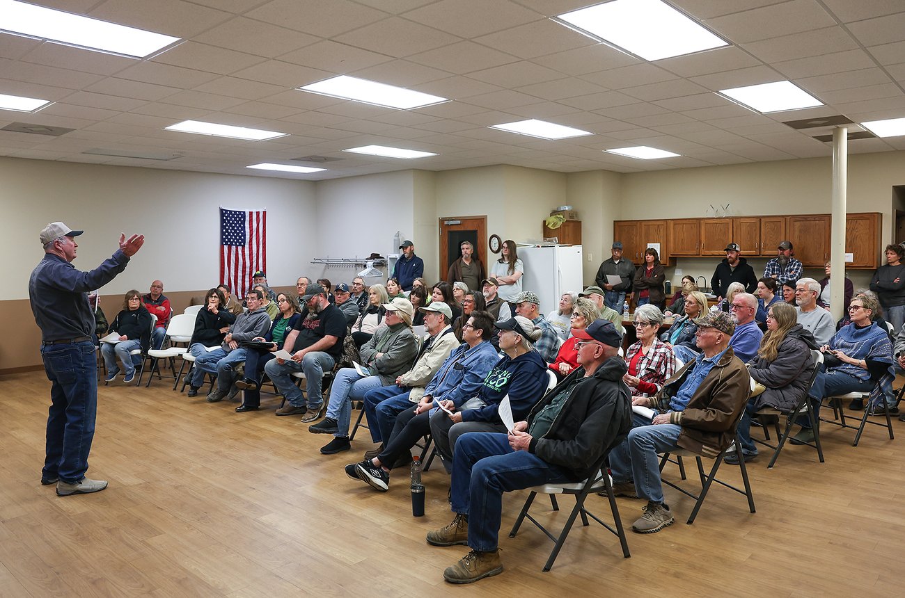 A crowd of people sitting in a social hall listen to a man speaking at the front of the room. All of them are dressed casually, with most wearing jeans, boots or sneakers, and light jackets. An American flag can be seen hanging on a wall in the background.