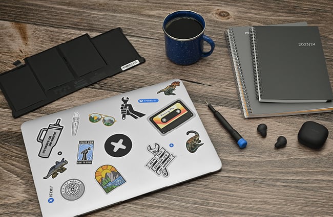 MacBook with stickers on a desk with a replacement battery, earbuds, and notebooks.