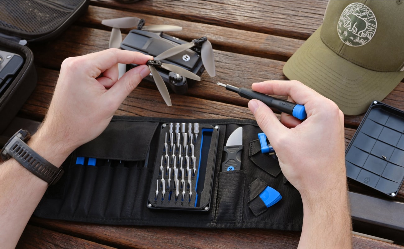 Hands repair a small drone on a wooden outdoor table, with an open iFixit toolkit laid out beneath the device and tools arranged for quick access.