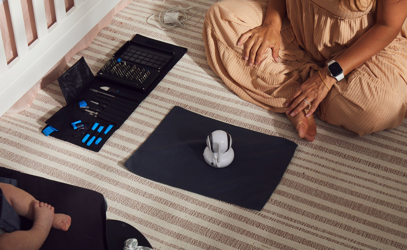 A mother sitting on the floor about to repair a baby monitor , Pro Tech® Toolkit open beside her and an infant's legs visibile in baby seat across from her.