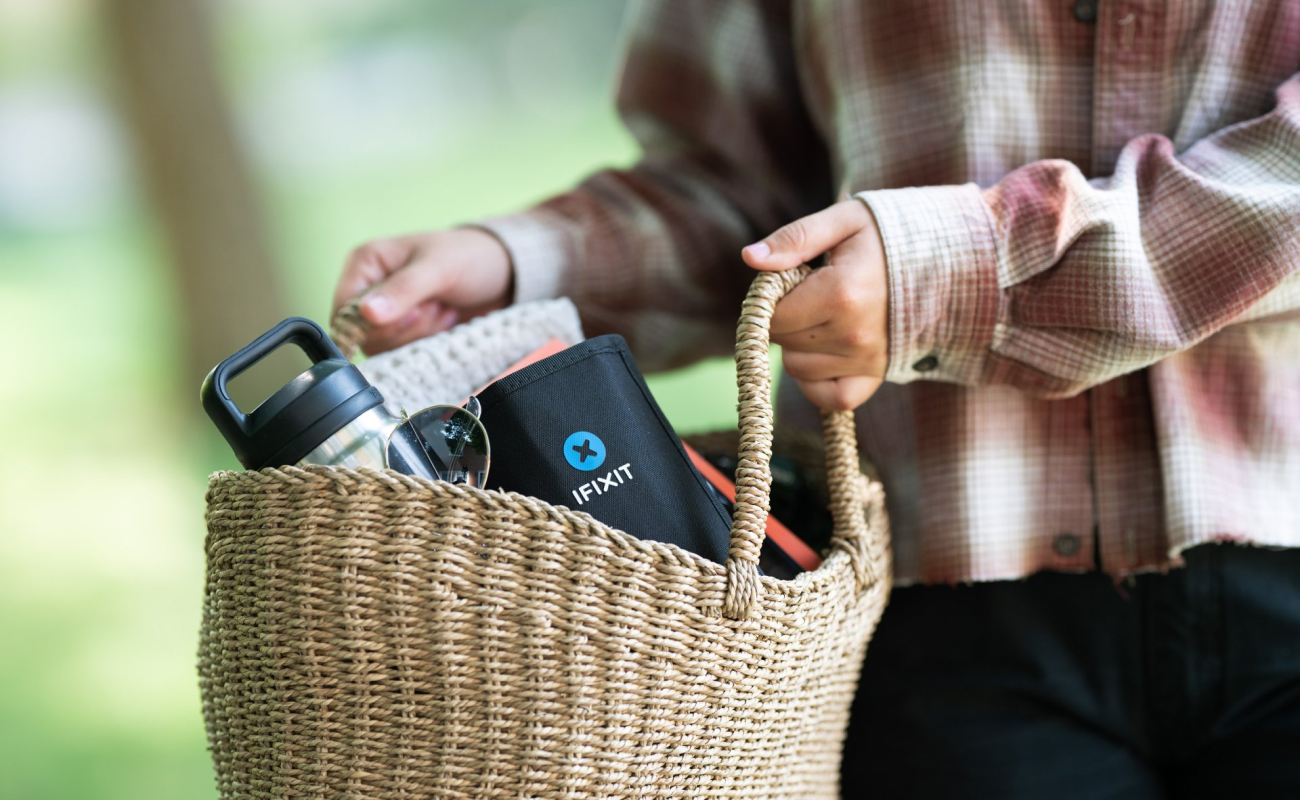A woman carries a woven basket outdoors with an iFixit toolkit, sunglasses, and a water bottle visible inside. The close-up composition keeps the basket and toolkit in focus against a softly blurred green background.
