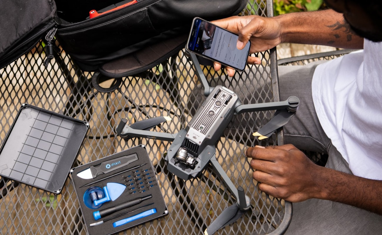 A person repairs or inspects a partially disassembled drone on an outdoor metal table, with an iFixit toolkit open nearby and a phone showing repair information. The composition is shot from above, focusing on the workspace, tools, hands, and drone.
