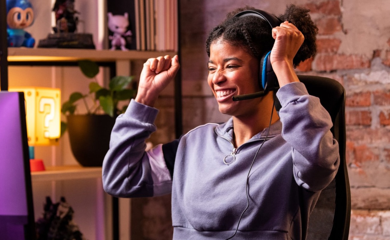 A smiling woman wearing a gaming headset raises both arms in celebration of a successful
repair while sitting at a desk in a warm, softly lit gaming setup.