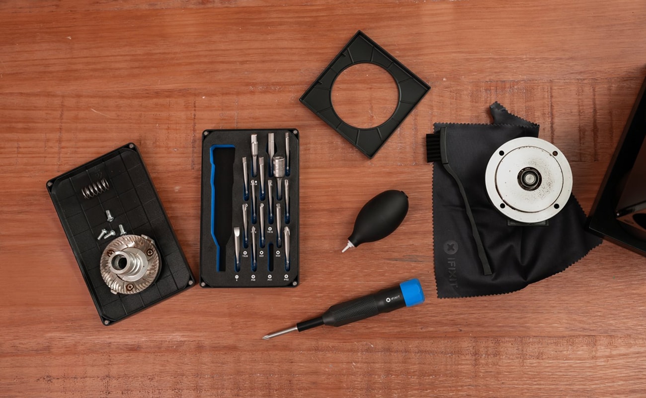 Overhead lifestyle image showing a coffee grinder repair setup on a wooden table, with an iFixit screwdriver, bit kit, air blower, cleaning cloth, screws, casing parts, and grinder components arranged around the workspace.