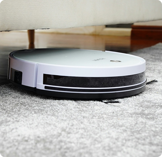 A white robot vacuum sits partially under a sofa on a light gray carpet, with a wooden floor and furniture details visible in the background. The low-angle composition focuses on the vacuum in the foreground as it navigates along the edge of the rug.