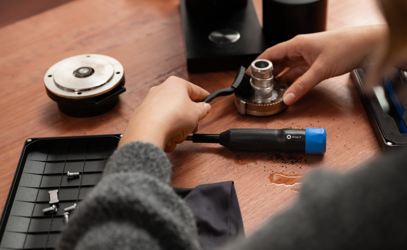 Close-up lifestyle photo of a person cleaning parts of a disassembled coffee grinder on a wooden table, with an iFixit Megalodon driver, bit set, screws, cloth, and brush nearby. The composition focuses on the hands working in the center, surrounded by repair tools and separated grinder components.