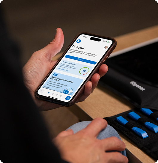 A person holds a smartphone showing the iFixit app interface next to an open tool roll with blue-handled tools on a wooden table. The close-up composition centers the phone screen in the hand, with the repair tools partially visible on the right.