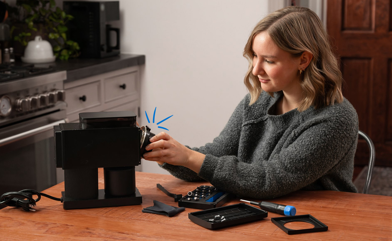 A woman repairs a black coffee machine at a wooden table, using driver from an open iFixit Megalodon Bit Kit while a blue graphic highlights “16 extended bits.” The composition places the repair scene in the center, with the bit kit and disassembled parts arranged in the foreground.