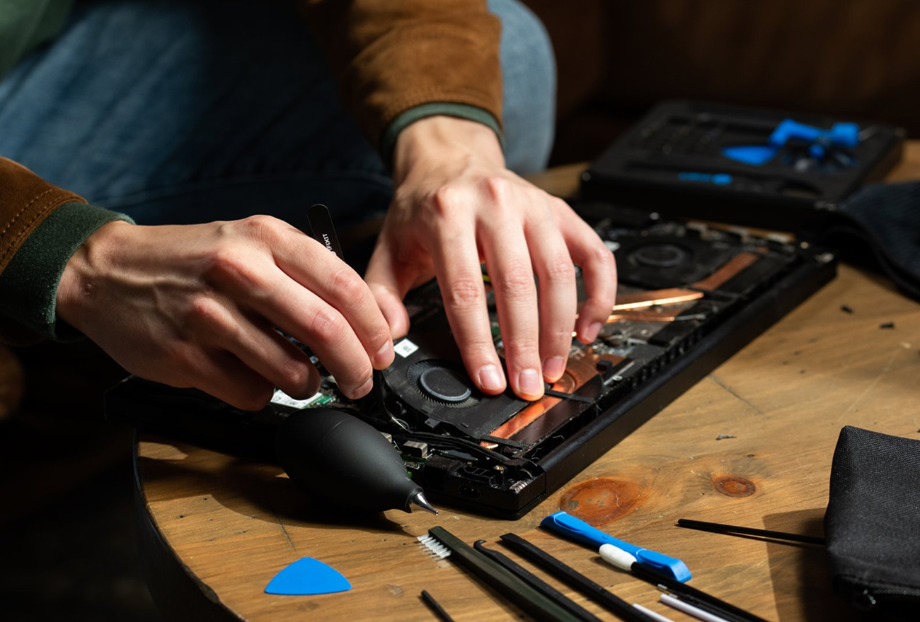 Close-up of a person using tweezers to repair an open laptop on a wooden table. The exposed cooling system is surrounded by blue opening tools, brushes, and other repair equipment.