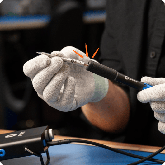 Close-up of gloved hands holding an iFixit Smart Soldering Iron with a soldering tip attached, positioned horizontally above a blue work mat.