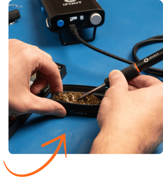 Close-up of hands cleaning the tip of an iFixit Smart Soldering Iron in a brass-wire tip cleaner on a blue work mat, with the soldering station visible in the background.