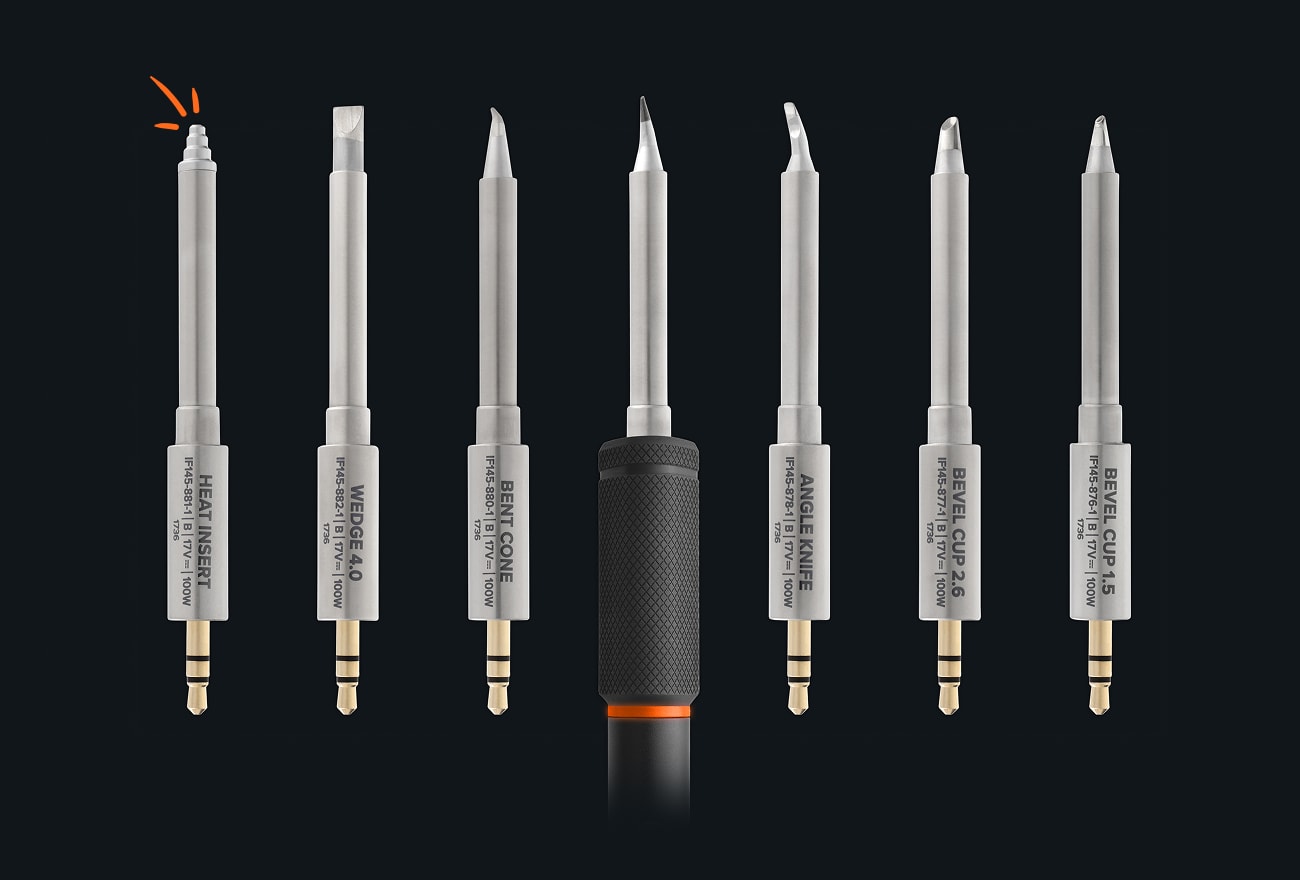 Seven interchangeable soldering tips are arranged vertically in a row on a dark background, with an iFixit Smart Soldering Iron centered among them.