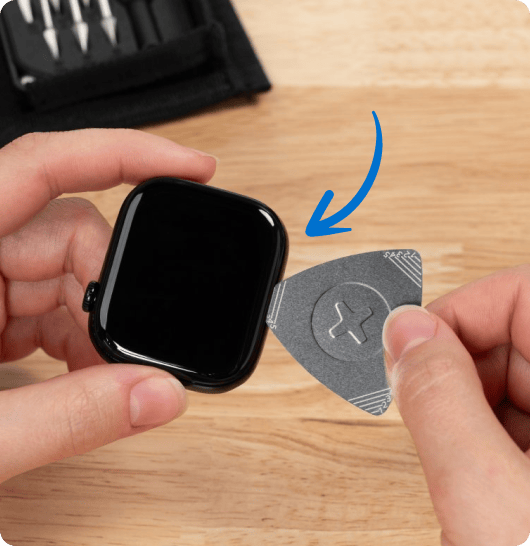 Two hands hold a smartwatch and a metal opening pick above a wooden table. A curved blue arrow points from the pick toward the edge of the watch.