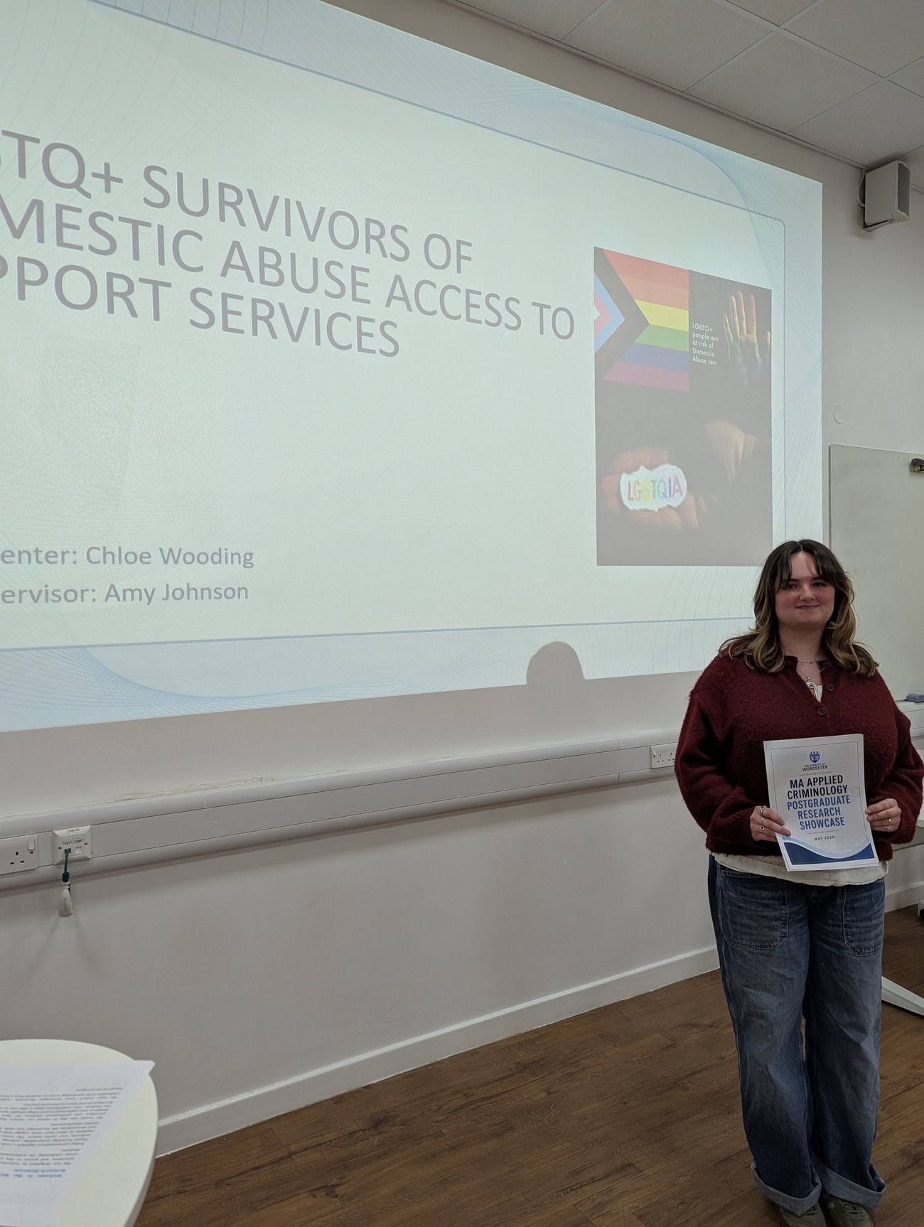 A young woman stands at the front of a presentation room holding a certificate from the MA Applied Criminology Postgraduate Research Showcase. Behind her, a projected presentation slide discusses LGBTQ+ survivors of domestic abuse and access to support services, featuring LGBTQ+ pride imagery. The woman smiles at the camera while standing beside the large projection screen.