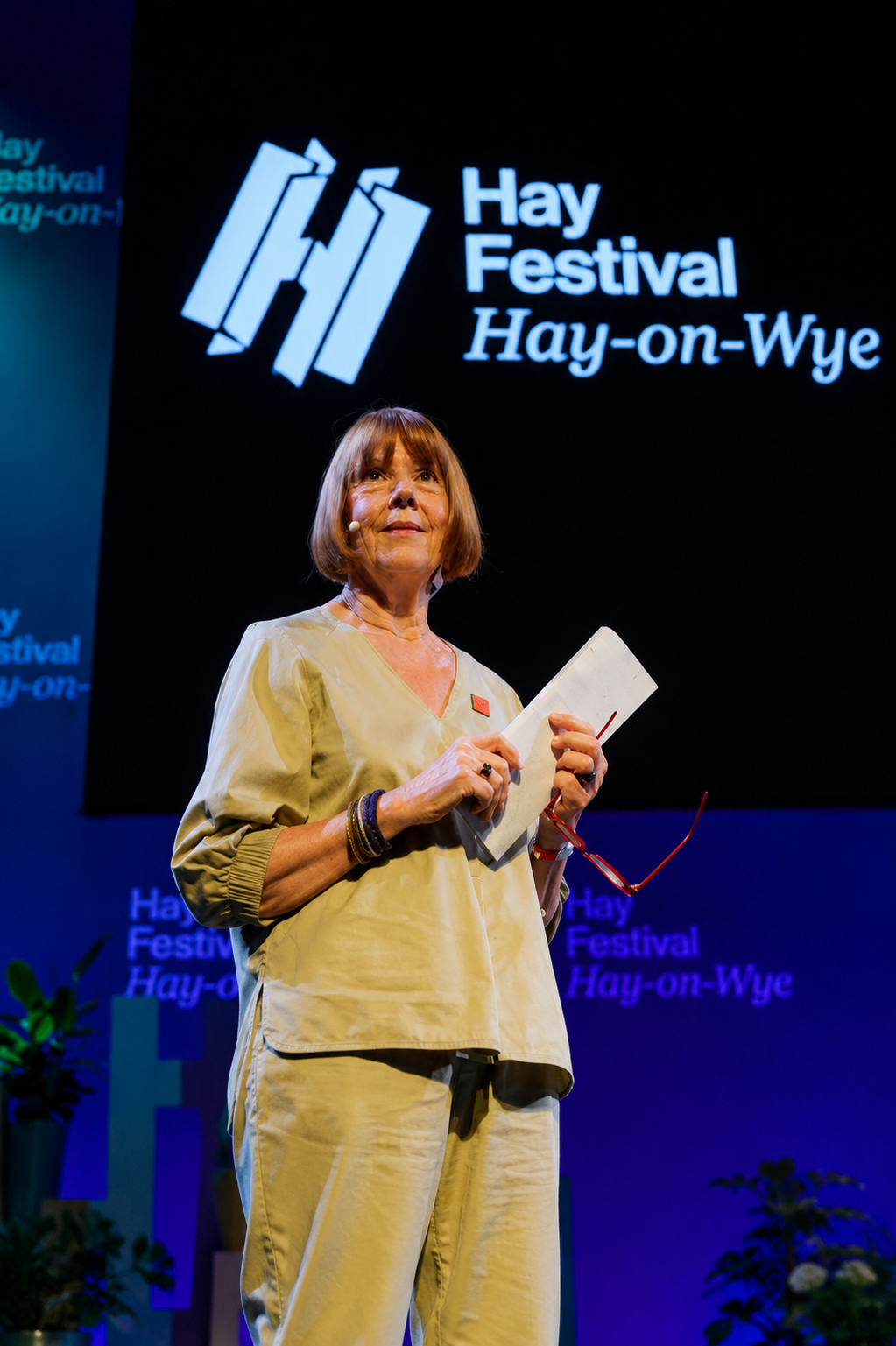A woman stands on stage at the Hay Festival in Hay-on-Wye, holding notes while addressing the audience. Behind her, a large screen displays the Hay Festival logo and name. The stage is lit with blue and purple lighting, and the woman is positioned in front of the backdrop, appearing to speak during a festival event. The woman is Gisèle Pelicot.